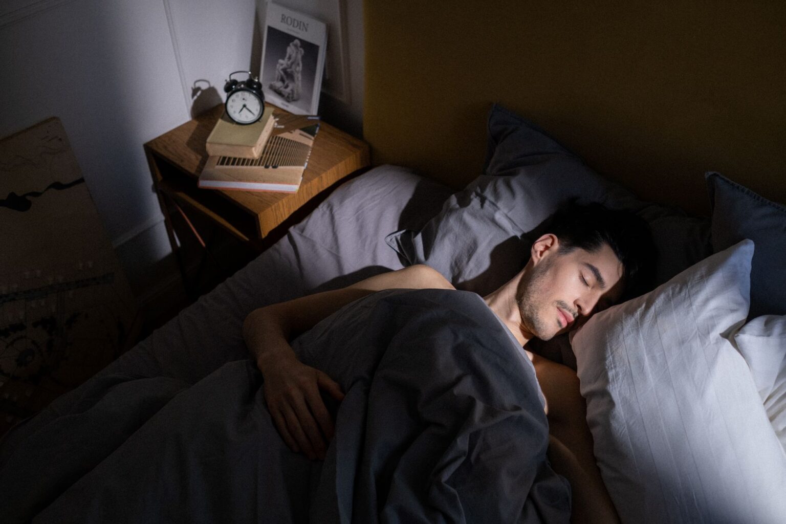 Mężczyzna śpi spokojnie w łóżku pod szarą kołdrą, a obok na stoliku nocnym stoi budzik i książki